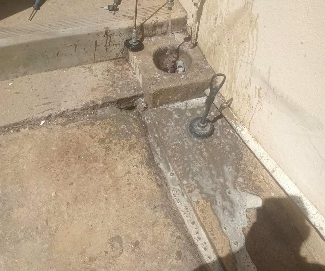 A blocked outside gully at the foot of a wall, drain rods and a rubber plunger head in use, soapy waste water spread across the concrete