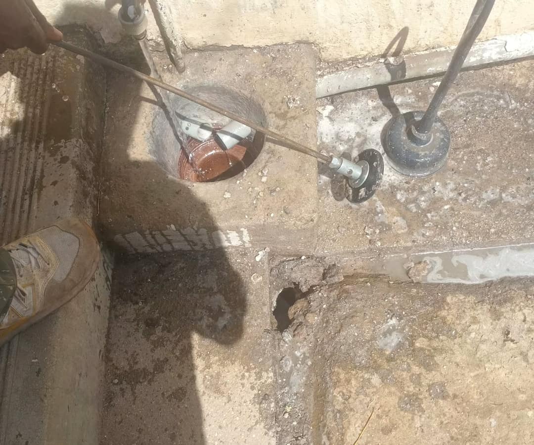 A plumber holding drain rods with a scraper disc beside a clay yard gully, water running into the gully and a rubber plunger head on the next rod